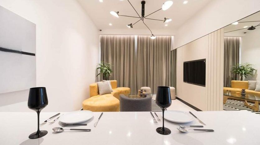 Une salle à manger moderne de l'Aria Résidence Dubai comprenant une table en marbre blanc avec des assiettes et des verres noirs, flanquée d'un canapé jaune moelleux et de chaises grises, avec un élégant luminaire suspendu et un