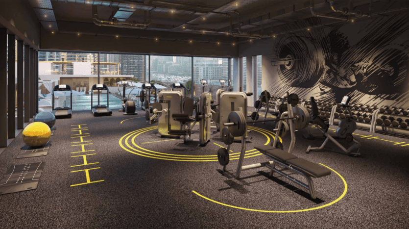 Intérieur de salle de sport moderne avec divers équipements d'exercice, notamment des tapis roulants, des poids et des appareils de résistance. Le sol est marqué de lignes jaunes et les grandes fenêtres du Studio One Dubai Marina montrent un horizon urbain à l'extérieur.