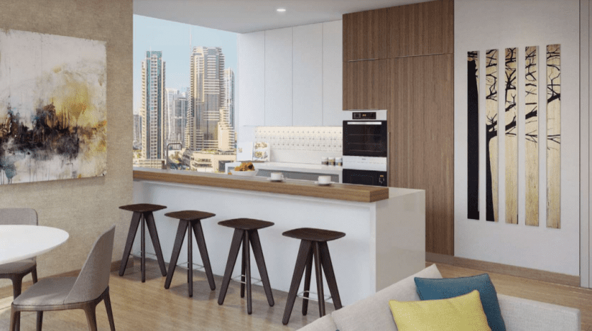 Intérieur de cuisine moderne comprenant un bar de petit-déjeuner avec des tabourets, des appareils électroménagers intégrés et de grandes fenêtres donnant sur le paysage urbain de Jumeirah Living Marina Gate. La décoration comprend de l'art abstrait et une salle à manger minimaliste.