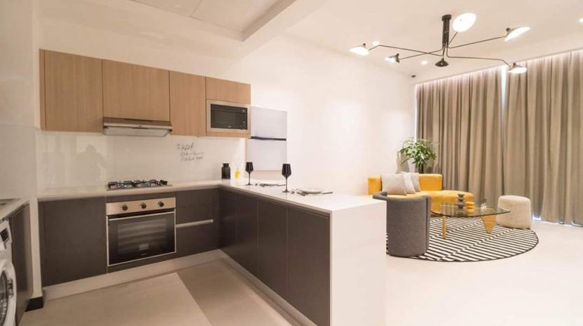 Une cuisine moderne avec des armoires grises, des accents en bois et des appareils électroménagers intégrés à l'Aria Residence Dubai. Le salon adjacent comprend un fauteuil jaune, un canapé et un tapis rayé. Éclairage et rideaux lumineux