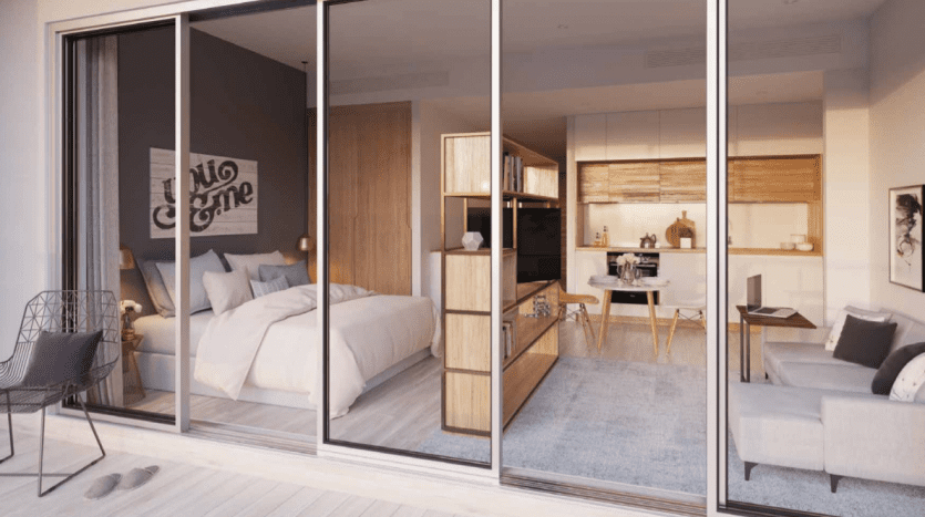 Intérieur moderne d'un studio dans la marina de Dubaï, vu à travers de grandes portes coulissantes en verre, comprenant une chambre confortable avec des œuvres d'art, une étagère en bois menant à un espace de vie avec un canapé gris et une cuisine dans le