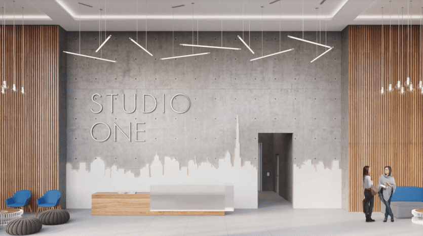 Lobby moderne et spacieux avec le lettrage « Studio One Dubai Marina » sur un mur gris, des panneaux en bois, des meubles minimalistes, des sièges colorés et deux personnes discutant avec désinvolture près d'un bureau de réception.