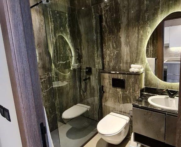 Une salle de bain moderne avec des murs en marbre foncé avec de grands miroirs, une douche à l'italienne, des toilettes blanches et un lavabo joya blanca. Une serviette blanche est suspendue à côté du lavabo et le sol est