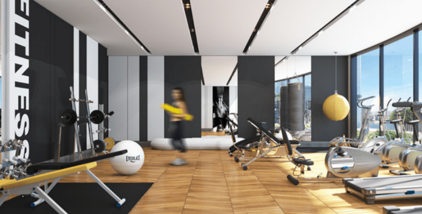 Une salle de sport moderne et bien équipée avec de grandes fenêtres dans la Nobles Tower, offrant une variété d'équipements de fitness comme des tapis roulants et des poids, et une silhouette floue d'une personne qui la traverse.