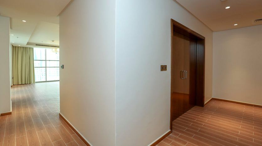 Intérieur d'appartement moderne vide dans la tour Amna, dans la ville d'Al Habtoor, avec parquet, murs blancs et porte en bois menant à une autre pièce ; la lumière naturelle traverse les fenêtres avec des rideaux.
