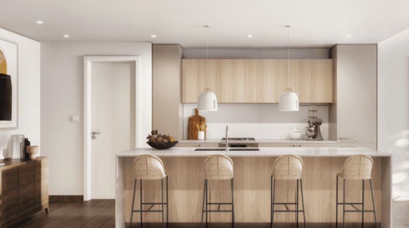 Cuisine moderne avec armoires en bois, comptoirs blancs, îlot central avec tabourets de bar et suspensions Sloane. Une porte à gauche et des appareils de cuisine sont visibles.