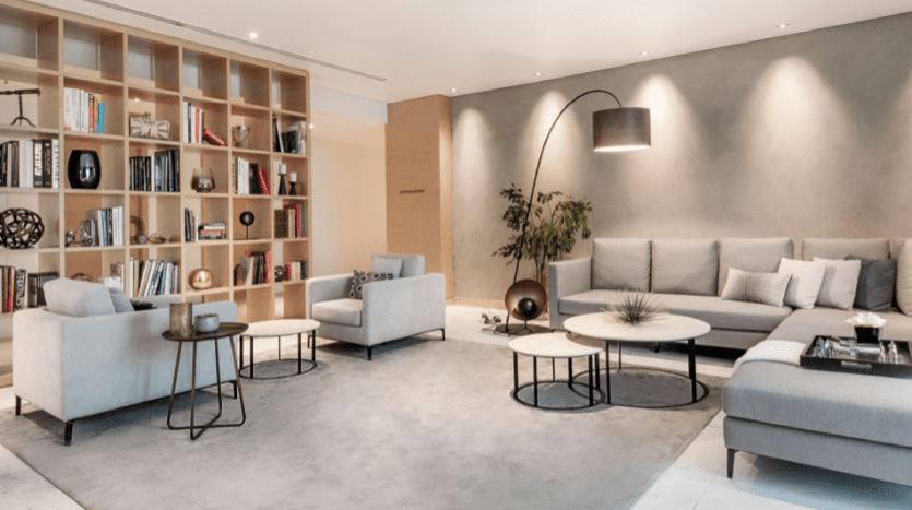Salon moderne comprenant un grand canapé sectionnel gris, plusieurs tables basses rondes, une bibliothèque complète et un lampadaire unique, avec un design minimaliste élégant adapté à l'immobilier Dubaï.