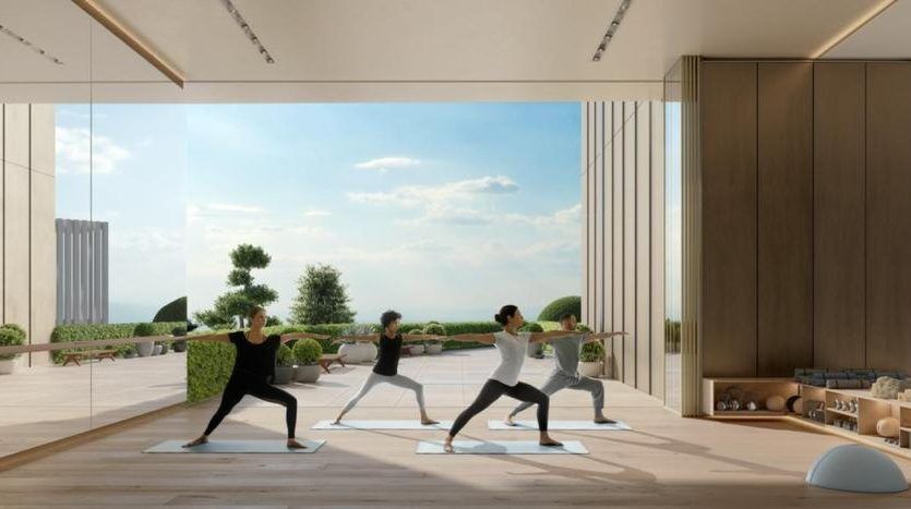 Trois personnes pratiquant le yoga dans une salle spacieuse et ensoleillée dotée de grandes fenêtres donnant sur un paysage extérieur serein à Dubaï. La chambre dispose de parquet et d'une décoration minimaliste.