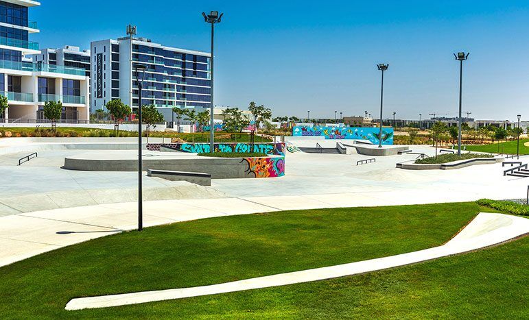 Un skatepark extérieur animé avec une variété de rampes et de rails de skate, des graffitis colorés sur les barrières, avec des bâtiments modernes en arrière-plan sous un ciel bleu clair à Kiara Damas Hills.