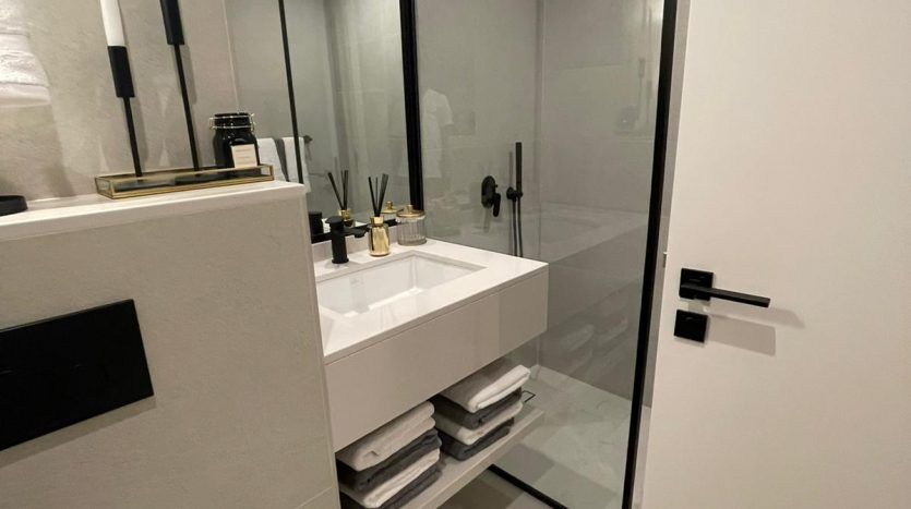 Une salle de bains moderne dans Prime Residency 3 comprenant une vanité blanche flottante avec un lavabo rectangulaire, des robinets noirs, un grand miroir et une cabine de douche en verre. La pièce est bien éclairée,