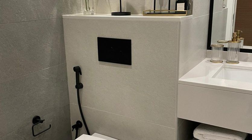 Une salle de bains moderne de Prime Residency 3 avec des carreaux gris comprend des toilettes murales blanches, un lavabo avec des robinets minimalistes et un vase avec des fleurs blanches. Luminaires noirs et serviettes blanches soigneusement empilées
