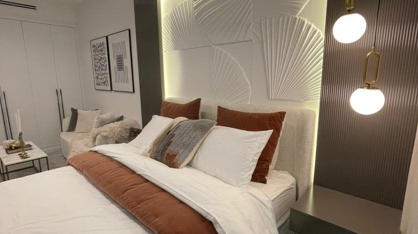 Une chambre moderne dans Prime Residency 3 avec un lit king-size soigneusement confectionné avec une literie blanche et marron. La pièce comprend des panneaux muraux artistiques avec rétroéclairage, une petite œuvre d'art encadrée et une suspension élégante.