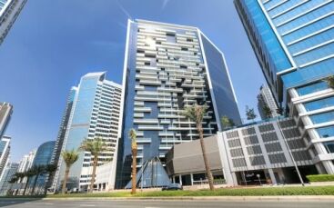 Paesaggio urbano moderno con alti grattacieli di vetro e Marquise Square Dubai con balconi. Palme rigogliose costeggiano la strada sotto un cielo azzurro e limpido.