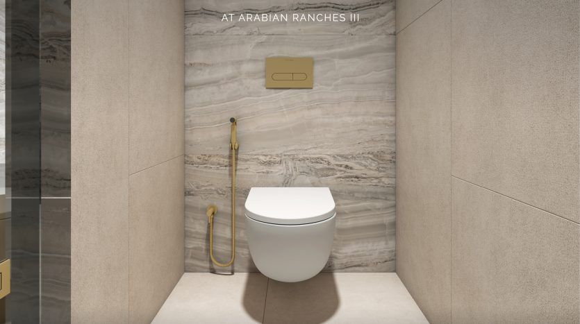 Intérieur de salle de bains luxueux avec toilettes murales modernes et luminaires dorés, mis en valeur par des murs en marbre texturé. L'espace est étiqueté "Elie Saab at Arabian Ranches".