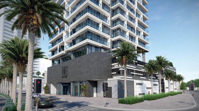 Immeuble moderne de grande hauteur avec balcons en verre, entouré de palmiers par une journée ensoleillée à Dubaï, comprenant des espaces commerciaux au rez-de-chaussée et une rue calme avec une voiture.