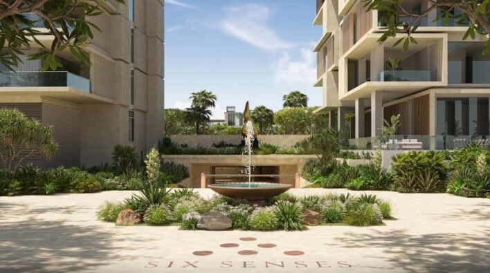 Une cour tranquille avec une fontaine d'eau circulaire au centre, entourée d'une verdure luxuriante et flanquée de bâtiments modernes à plusieurs étages sous un ciel dégagé. Le sol comporte le logo "Six Senses