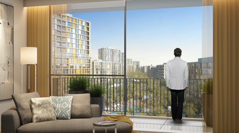 Un homme en chemise blanche regarde par une grande fenêtre dans un salon moderne, observant le paysage urbain de Dubaï. La pièce dispose d'un canapé élégant, d'un tapis décoratif et d'un éclairage élégant.
