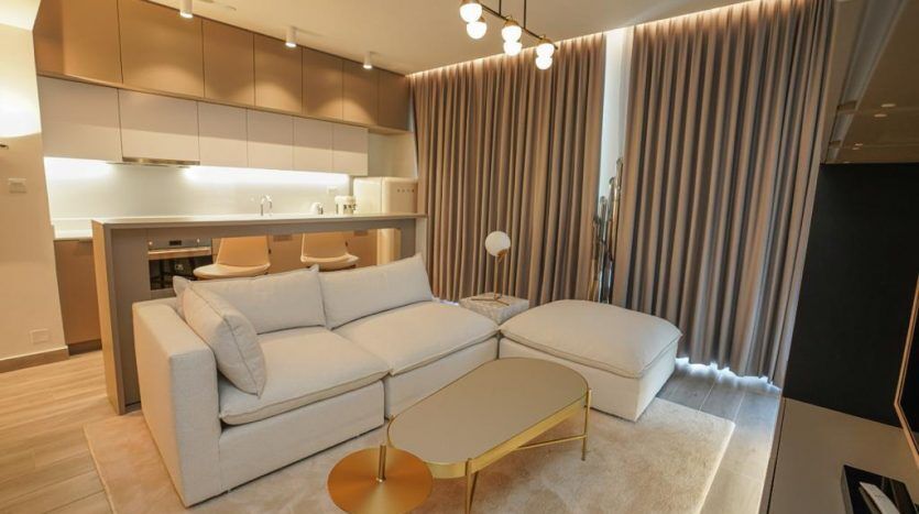 Un salon moderne dans une villa de Dubaï avec un canapé sectionnel blanc, une table basse dorée et une cuisine avec des armoires beiges en arrière-plan. Des rideaux de couleur claire et un éclairage chaleureux créent une atmosphère chaleureuse