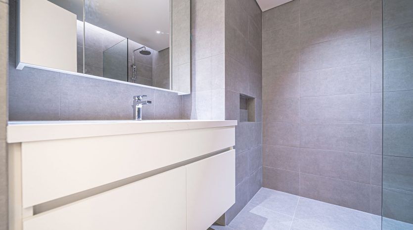 Une salle de bains moderne dans un appartement de Dubaï avec des carreaux gris clair, comprenant une vasque blanche avec un lavabo, un grand miroir et une étagère dissimulée. L'espace est bien éclairé et dispose d'un espace propre