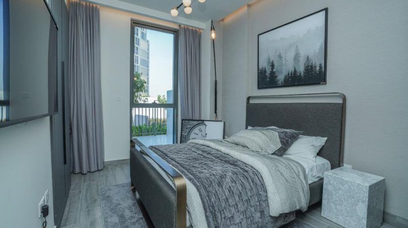 Une chambre moderne dans une villa de Dubaï comprenant un lit double avec une literie grise, deux photos de paysages de montagne encadrées au mur et un balcon avec vue sur des immeubles de grande hauteur. Rideaux et suspensions gris clair