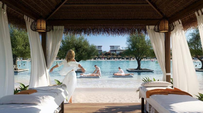 Une femme vêtue d'une robe blanche profite d'une vue panoramique sur un lagon relaxant avec des gens flottant sur des structures gonflables, depuis une cabane luxueuse ornée de lits moelleux et d'un décor naturel dans une villa exclusive de Dubaï.