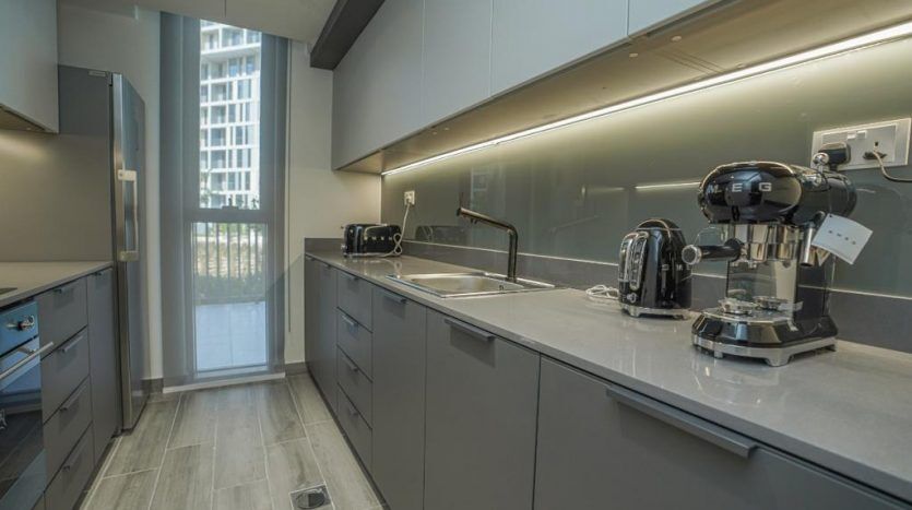 Intérieur de cuisine moderne dans un appartement de Dubaï comprenant des armoires grises élégantes, des appareils électroménagers intégrés et une machine à café, avec de la lumière naturelle provenant d'une fenêtre avec vue sur la ville.