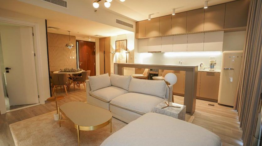 Salon d'appartement moderne à Dubaï avec un canapé beige, des tables basses dorées, une cuisine intégrée et un coin repas visible en arrière-plan. Un éclairage chaleureux et une ambiance cosy.