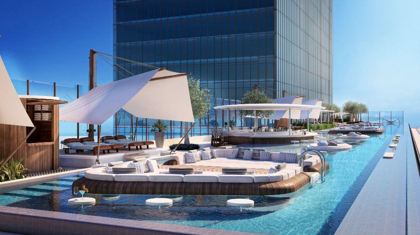 Espace luxueux au bord de la piscine dans un hôtel moderne de Dubaï, doté de sièges luxueux, de cabanes et de parasols avec vue sur l'océan, adjacent à un élégant bâtiment de grande hauteur.