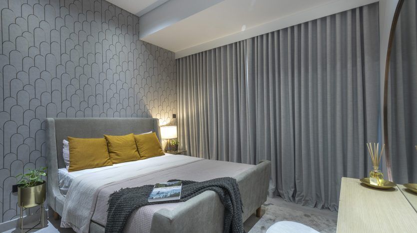 Une chambre moderne dans un appartement de Dubaï avec un grand lit recouvert d'oreillers gris et jaunes, du papier peint texturé, de lourds rideaux gris et un éclairage tamisé. Les objets de décoration comprennent des livres et une petite lampe.