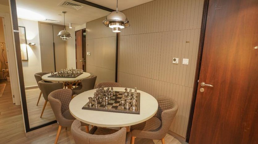 Une pièce confortable dans un appartement de Dubaï comprend deux tables rondes, chacune entourée de chaises. Une table contient un jeu d’échecs prêt à jouer. Un éclairage chaleureux et un mélange de murs en bois et gris texturés créent une