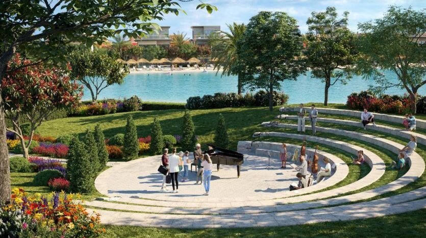Les gens se rassemblent dans un jardin luxuriant avec une zone centrale pavée ronde dotée d'un piano, entourée de parterres de fleurs colorés et d'une vue panoramique sur un lac en arrière-plan d'un immobilier Dubaï.