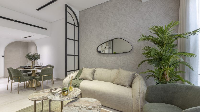 Un salon moderne avec un canapé beige, deux fauteuils verts et une table basse en marbre. Derrière, un espace salle à manger de l'appartement Dubai sous une ouverture cintrée. Grandes plantes et papier peint géométrique