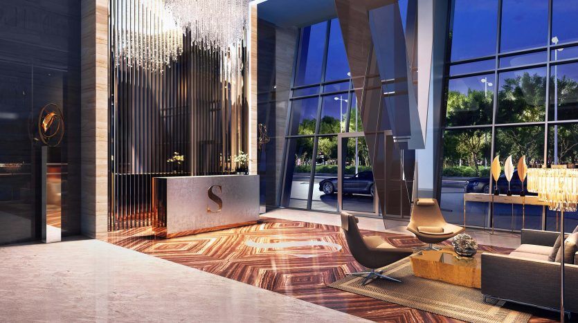 Le soir, le hall d'entrée luxueux de l'hôtel présente une décoration moderne avec de hauts plafonds, un éclairage élégant, de grandes fenêtres et un coin salon élégant. L'entrée du Sterling Dubai affiche un logo « S » bien visible sur le