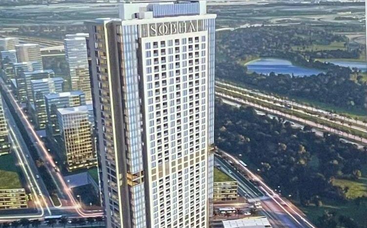 Vue aérienne d'un grand gratte-ciel, étiqueté « Skoda », intégré à un complexe de bâtiments inférieurs, adossé à l'horizon de la ville avec de nombreux autres gratte-ciel et un parc verdoyant à Dubaï