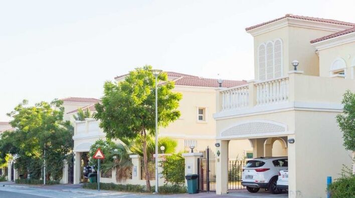 Une rue de banlieue calme bordée de maisons modernes à deux étages dotées de balcons et de portes cintrées, d'arbres verts luxuriants et de voitures garées sous un ciel clair se transforme en un quartier chic de Dubaï où les investisseurs