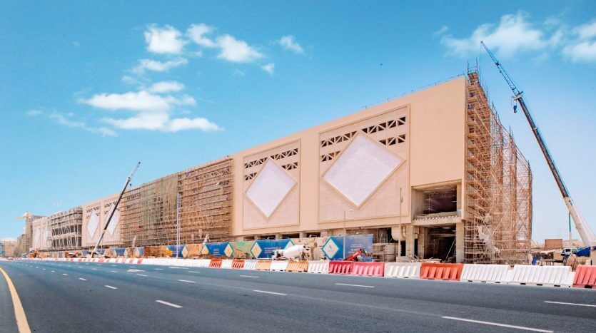 Un grand chantier de construction le long d'une autoroute avec des grues et des échafaudages autour d'un bâtiment beige partiellement achevé avec de grands motifs en forme de losange sur sa façade sous un ciel bleu clair à Dubaï.