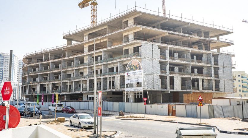 Un chantier de construction à plusieurs niveaux d'un grand bâtiment avec une signalisation claire pour les « développeurs sûrs » et la « villa Dubaï ». Une grue est visible sur un ciel clair, avec des véhicules et des barrières de construction à proximité.