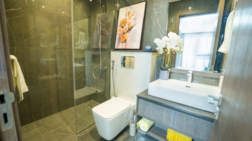 Salle de bains moderne comprenant une cabine de douche en verre, des toilettes blanches et un lavabo surmonté d'un grand miroir sur lequel est exposée une peinture florale. Il y a des orchidées sur le comptoir de l'évier et des carreaux gris dans toute la villa à Dubaï