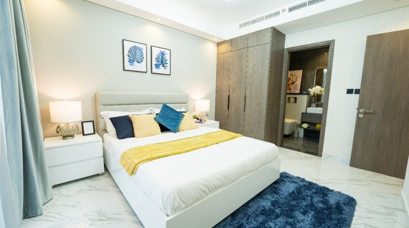 Une chambre moderne et élégante dans une villa de Dubaï avec un lit soigneusement fait, avec une literie blanche et jaune. Les accents bleus sont visibles à travers un tapis et une décoration murale. Boiseries et porte menant à