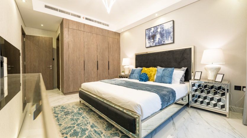 Chambre moderne dans une villa de Dubaï comprenant un grand lit avec une literie bleue et blanche, une tête de lit en bois sombre, des tables de nuit élégantes avec lampes, un tapis bleu et des armoires en bois, éclairées par le plafond