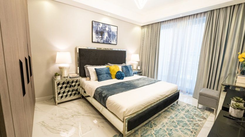 Chambre moderne dans une villa à Dubaï avec un grand lit doté d'une tête de lit capitonnée, d'une literie bleue et blanche et d'oreillers jaunes. La décoration comprend une commode, des lampes, un tapis,