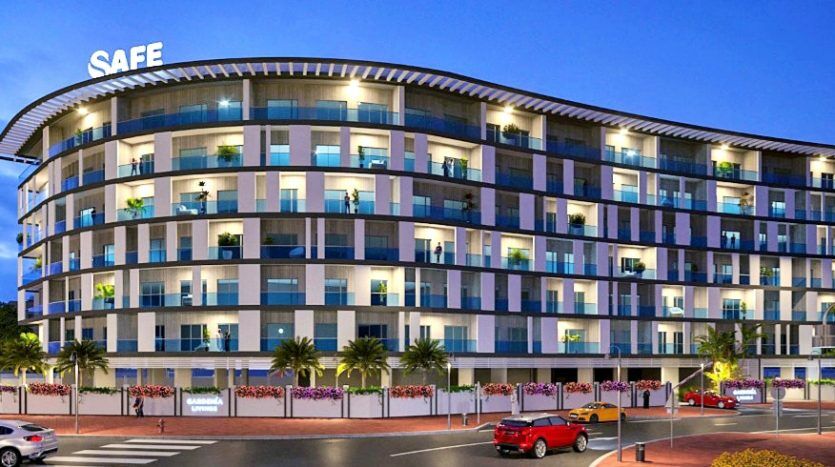 Un bâtiment moderne et incurvé de plusieurs étages à Dubaï, illuminé au crépuscule, doté de balcons avec des balustrades en verre et des fleurs éclatantes. Le bâtiment est étiqueté « sûr » en grosses lettres en haut. Les voitures sont