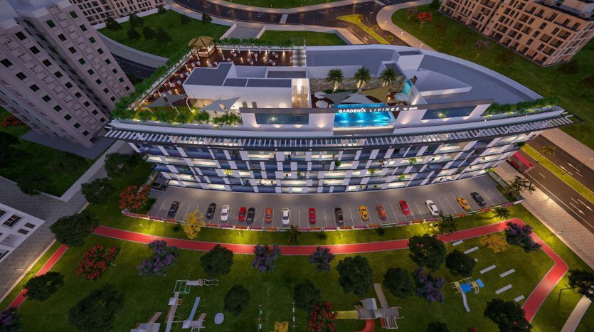 Vue aérienne d'un bâtiment moderne de plusieurs étages à Dubaï au crépuscule avec des intérieurs éclairés et des lumières extérieures. Les voitures sont garées à côté, entourées de jardins paysagers et d'allées.