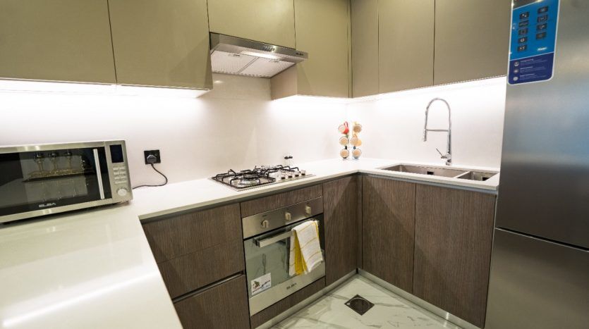 Une cuisine moderne dans un appartement de Dubaï avec des armoires en bois, un four micro-ondes intégré, un four, une cuisinière et un évier. Il y a une tasse décorative posée sur le comptoir et une commande numérique