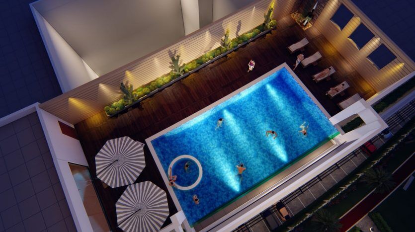 Vue aérienne d'un toit la nuit à Dubaï avec une grande piscine entourée de chaises longues, une terrasse en bois et des jardinières vertes, éclairées par un éclairage ambiant.