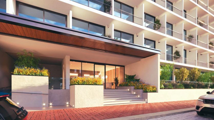 Entrée d'un immeuble d'appartements moderne à Dubaï avec de grandes portes vitrées, des marches éclairées et des balcons bordés de plantes. Voitures garées près de l’entrée sous un ciel coucher de soleil.