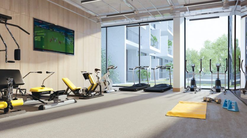 Une salle de sport moderne avec de grandes fenêtres, dotée de divers équipements d'exercice, notamment des tapis roulants, des vélos et des bancs de musculation, entourée d'une vue panoramique. Une télévision diffusant un match de sport est accrochée au mur de