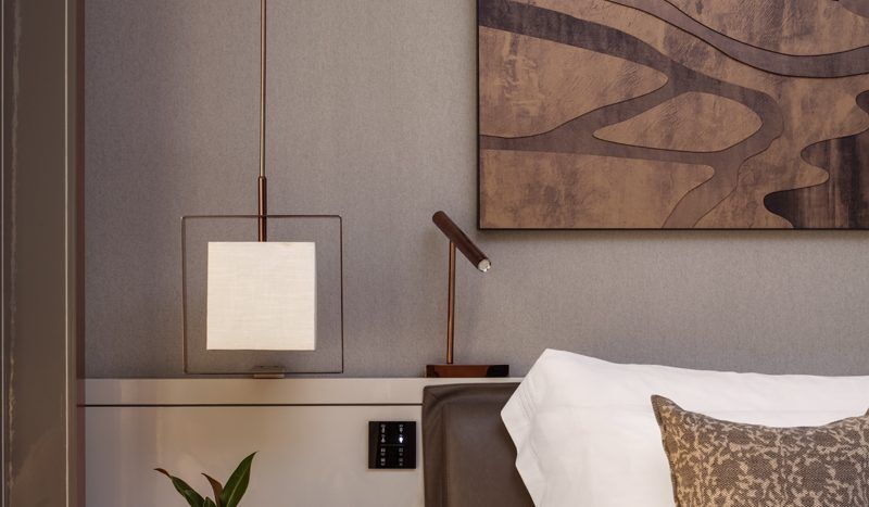 Un coin chambre confortable dans une propriété de l'immobilier Dubaï comprenant un lit soigneusement fait avec des draps blancs et des oreillers décoratifs, une table de chevet avec une lampe et une œuvre d'art abstraite en bois au mur.
