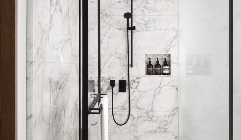 Douche de salle de bains moderne dans une villa de Dubaï avec des murs en marbre blanc, une porte vitrée et des accessoires noirs, dont une pomme de douche à effet pluie et un pulvérisateur à main. Une serviette est accrochée au mur.