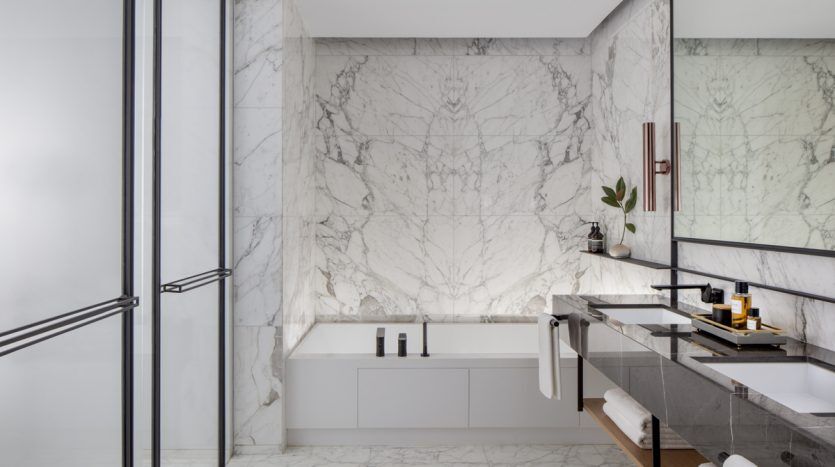 Salle de bains luxueuse dans un emplacement privilégié de l'immobilier à Dubaï, avec de grands murs et sols en marbre, une baignoire blanche autoportante, des armoires élégantes et d'élégants accents dorés sous un éclairage tamisé.